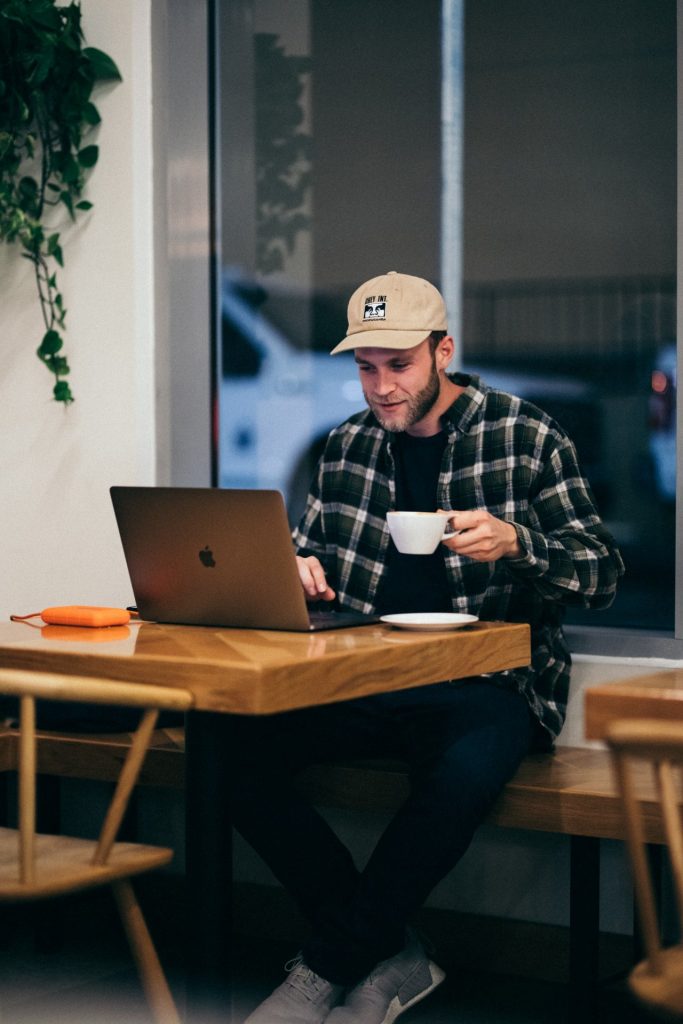 Jonge man achter zijn laptop aan het werk in een cafe met een kop koffie in zijn hand. Afbeelding is puur decoratief.
