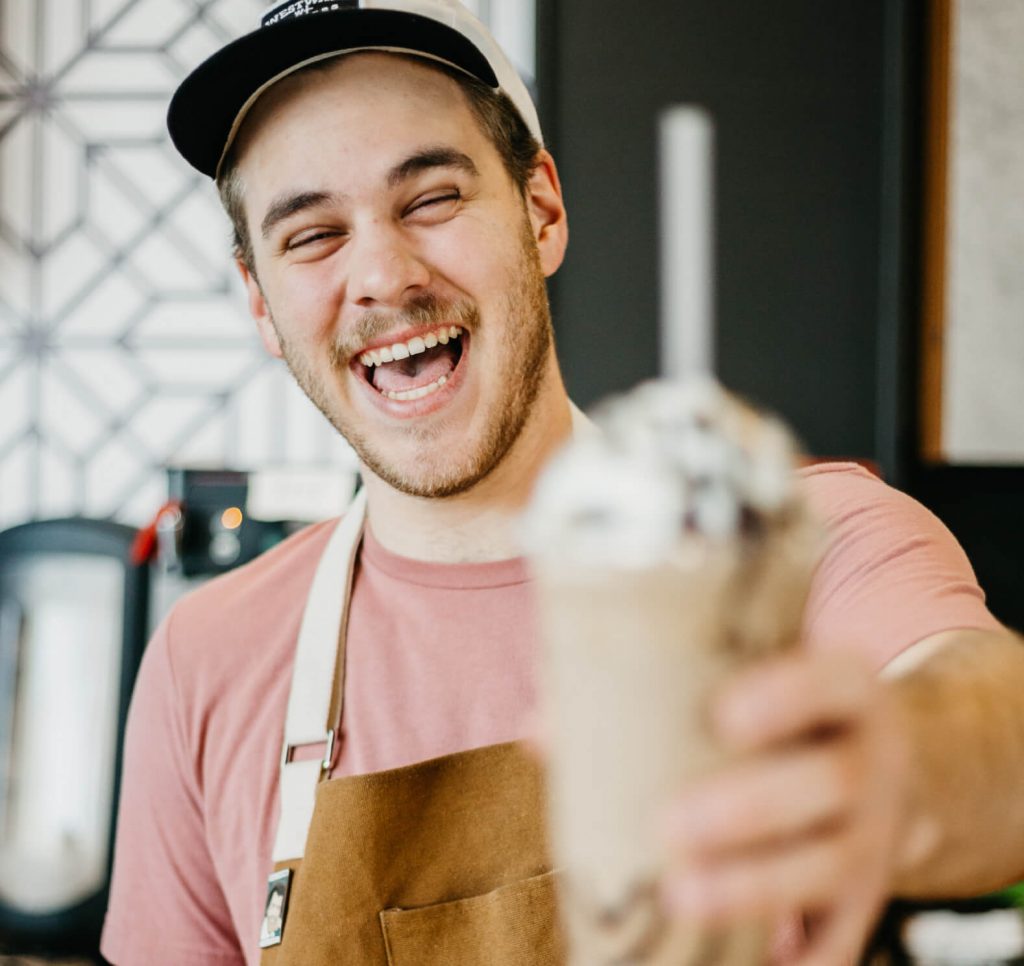 Een blije, blonde mannelijke barista met een baartje. Hij heeft een milkshake in zijn handen, een roze shirt en een schort aan, en een pet op zijn hoofd.